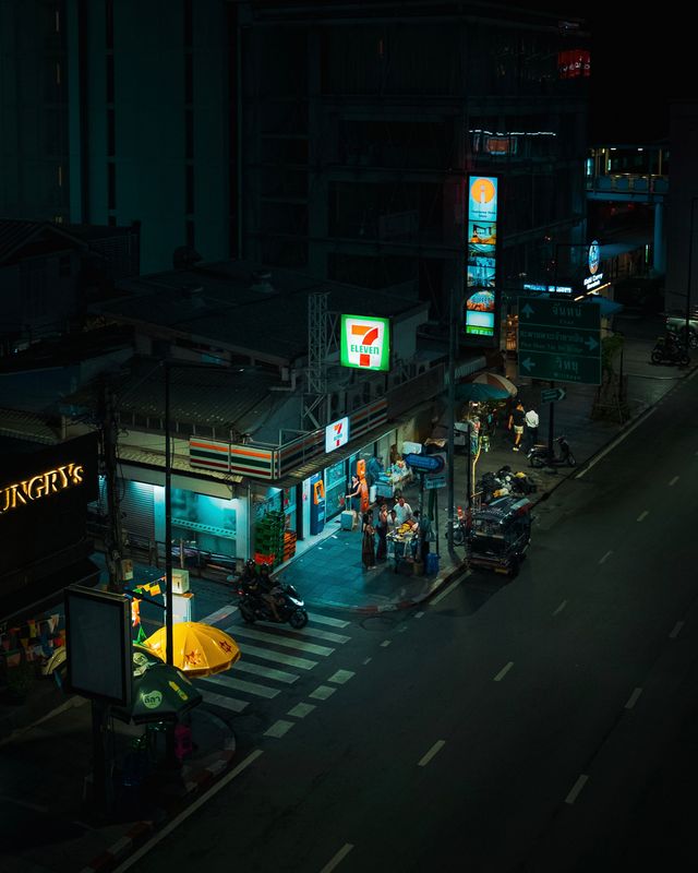 A quiet Bangkok street at night, lit by a convenience store and a pedestrian crossing.