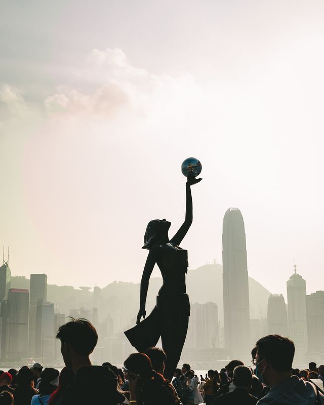 A statue raising a ball silhouetted against the Hong Kong skyline in hazy afternoon light.