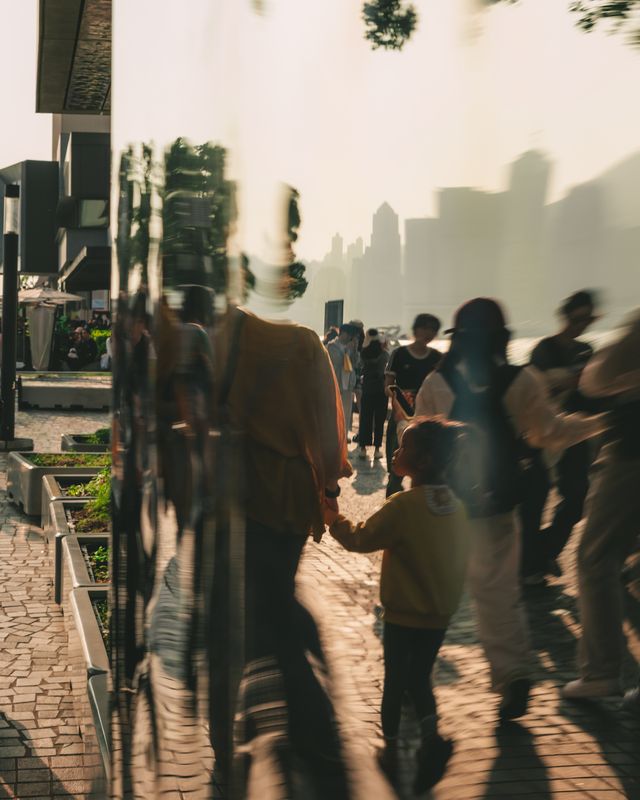 Commuters and a child walking through long afternoon shadows on a Hong Kong street.