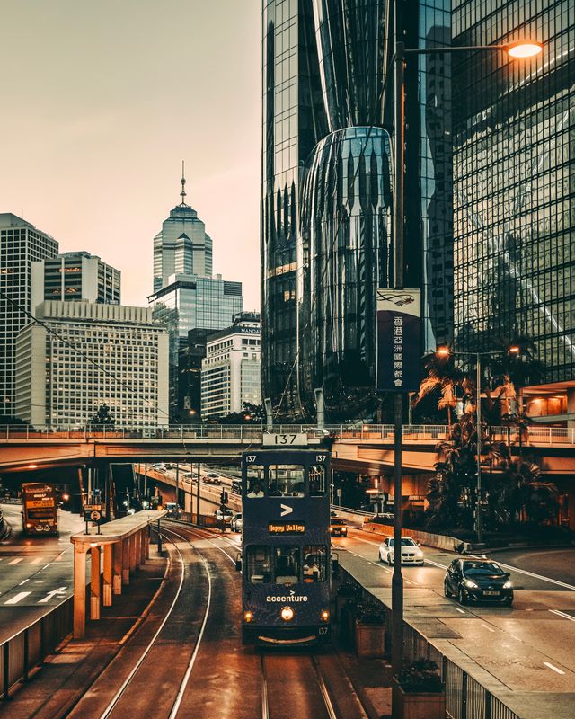A Hong Kong tram on a wide road at dusk, with skyscrapers reflected in the glass behind it.