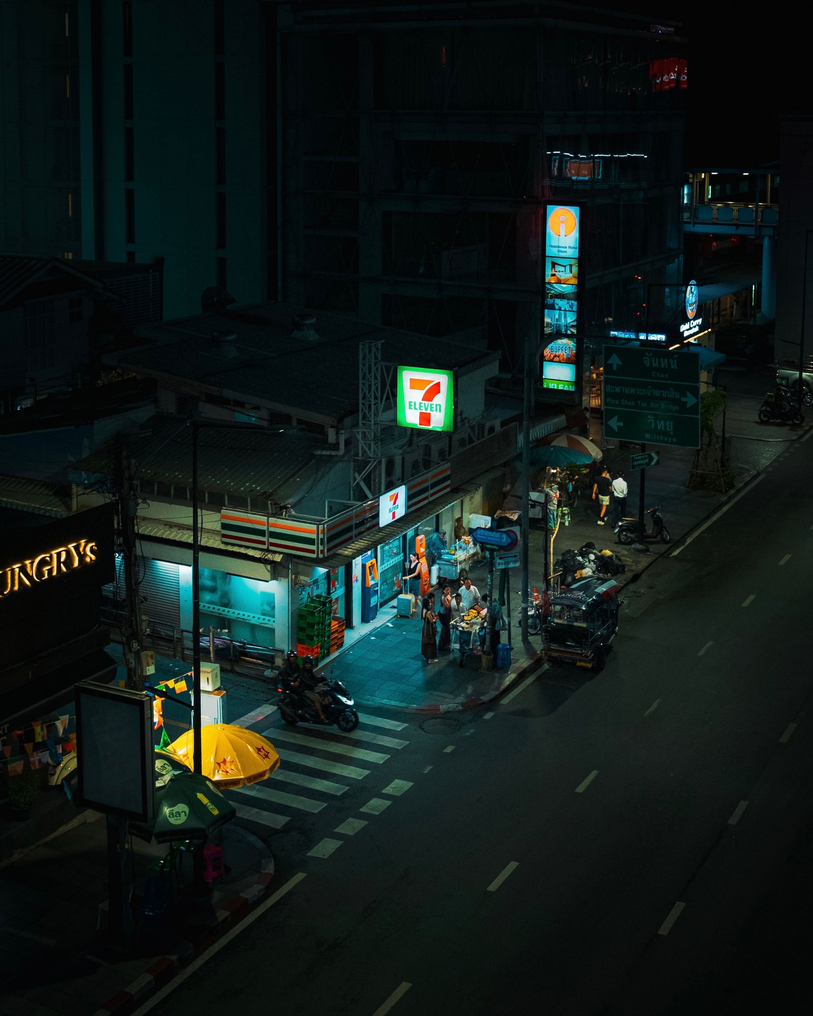 A quiet Bangkok street at night, lit by a convenience store and a pedestrian crossing.