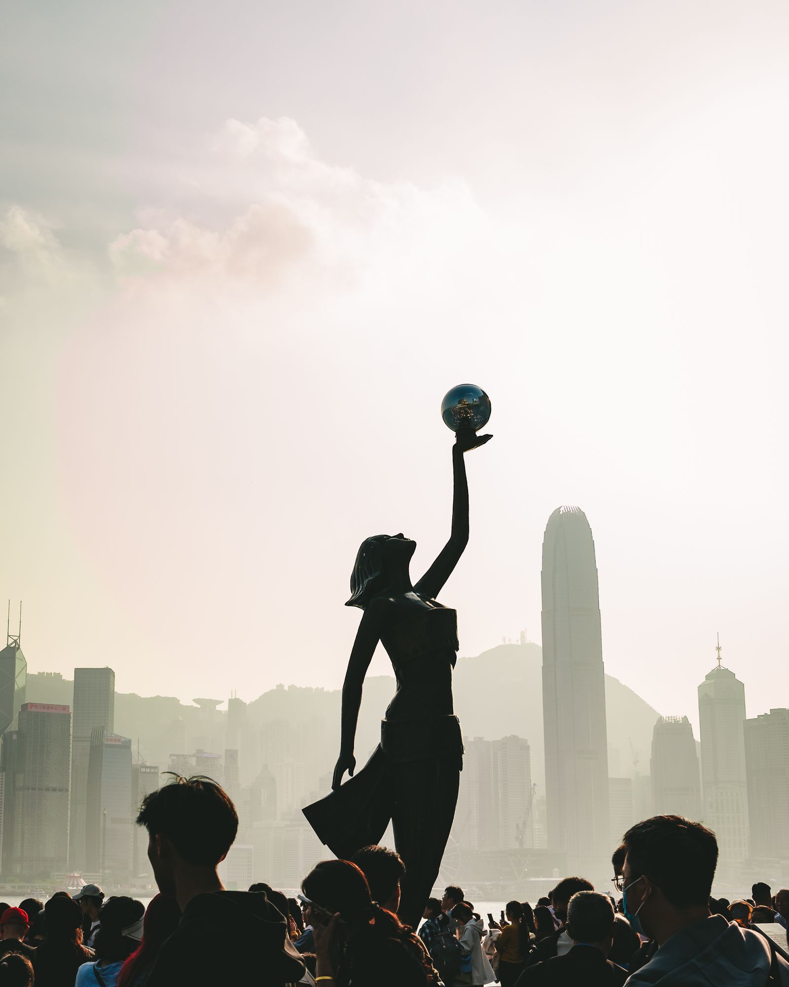 A statue raising a ball silhouetted against the Hong Kong skyline in hazy afternoon light.