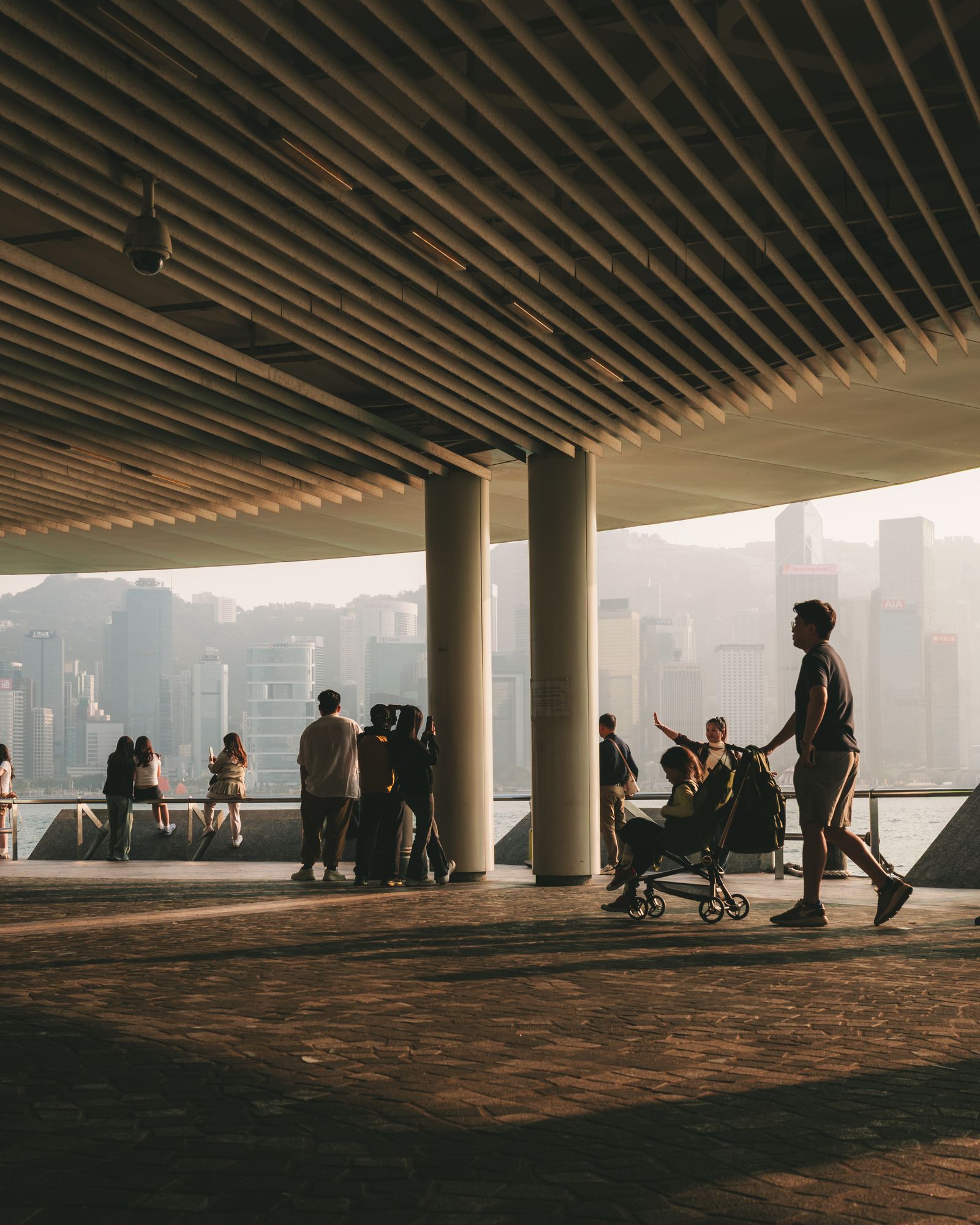 A long covered waterfront colonnade with people scattered across the sunlit floor and the harbour beyond.